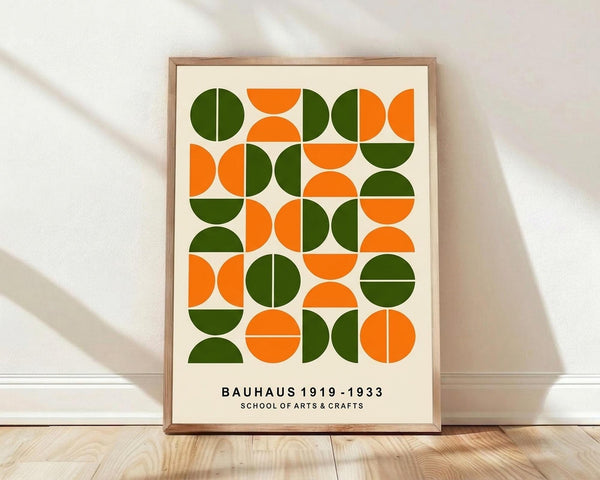 May include: Framed geometric art print featuring alternating green and orange circles and semicircles in a grid pattern on cream background, displayed in light wood frame leaning against white wall on blonde hardwood floor with soft natural lighting and shadows