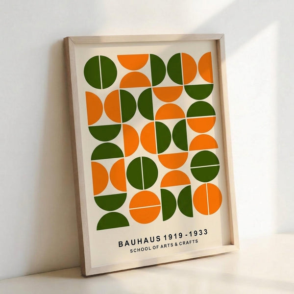 May include: Framed geometric poster with orange and green circular and semi-circular shapes arranged in a grid pattern on cream background, displayed in light wooden frame leaning against white wall. The design features bold mid-century modern style with "BAUHAUS 1919-1933 SCHOOL OF ARTS & CRAFTS" text at bottom.