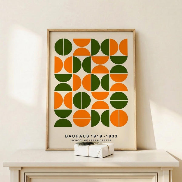 May include: Framed geometric poster with green and orange circles and semicircles arranged in a grid pattern on cream background, displayed on light wood console table next to small white gift box, against neutral wall with natural lighting