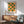 May include: Framed geometric poster with orange and green circles and semicircles in a grid pattern, displayed above a wooden sideboard with white ceramic lamp. Mid-century modern sage green armchair with wooden frame sits nearby on wooden floor. Dramatic window light creates shadows on white wall.