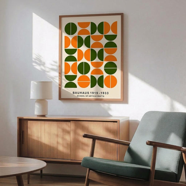 May include: Framed geometric poster with orange and green circles and semicircles in a grid pattern, displayed above a wooden sideboard with white ceramic lamp. Mid-century modern sage green armchair with wooden frame sits nearby on wooden floor. Dramatic window light creates shadows on white wall.