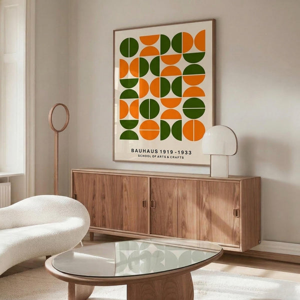 May include: Mid-century modern living room featuring a framed geometric poster with orange and green semicircles arranged in a grid pattern. The artwork hangs above a walnut wood credenza with sliding doors, next to a white curved lounge chair and glass-top oval coffee table. Minimalist decor with neutral walls and natural lighting.