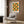 May include: Modern geometric poster with green and orange semicircles arranged in a grid pattern, displayed in light wood frame on wood paneled wall above cream upholstered armchair with patterned pillow. Brass floor lamp and side table with open book complete the minimalist reading nook scene with natural lighting from window.