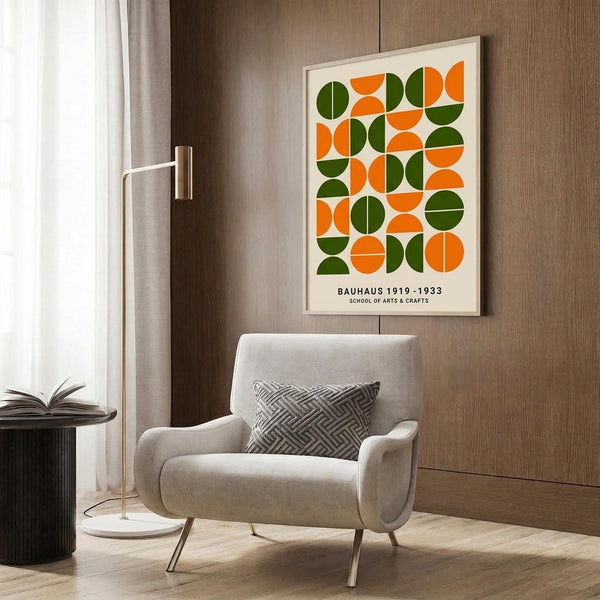 May include: Modern geometric poster with green and orange semicircles arranged in a grid pattern, displayed in light wood frame on wood paneled wall above cream upholstered armchair with patterned pillow. Brass floor lamp and side table with open book complete the minimalist reading nook scene with natural lighting from window.