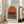 May include: Framed minimalist art print featuring a large orange circle above horizontal black stripes on cream background, displayed in light wood frame leaning against white wall next to beige sofa in bright living room with large windows and hardwood floors.