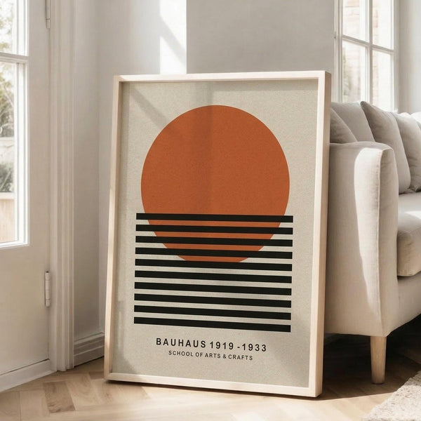 May include: Framed minimalist art print featuring a large orange circle above horizontal black stripes on cream background, displayed in light wood frame leaning against white wall next to beige sofa in bright living room with large windows and hardwood floors.