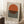 May include: Framed poster with orange semicircle above black and white horizontal stripes on cream background, leaning against white wall corner. Light wooden frame, displayed on white textured rug near wooden side table with white coffee cup and saucer. Soft natural lighting creates shadows on wall.