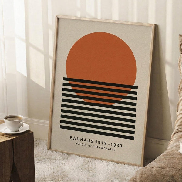 May include: Framed poster with orange semicircle above black and white horizontal stripes on cream background, leaning against white wall corner. Light wooden frame, displayed on white textured rug near wooden side table with white coffee cup and saucer. Soft natural lighting creates shadows on wall.