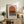 May include: Framed geometric art print with orange semicircle above black horizontal stripes on cream background, displayed on rustic wooden sideboard alongside fern plant, leather armchair, ceramic vase, and stacked books in warm neutral living room setting.
