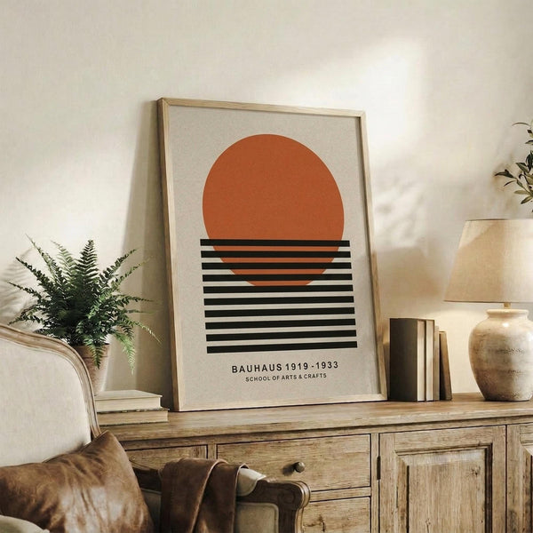 May include: Framed geometric art print with orange semicircle above black horizontal stripes on cream background, displayed on rustic wooden sideboard alongside fern plant, leather armchair, ceramic vase, and stacked books in warm neutral living room setting.