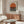 May include: Modern living room with walnut credenza displaying framed poster featuring orange sun above black horizontal stripes on cream background. White sculptural chair, glass coffee table, and copper floor lamp complete the mid-century inspired space with neutral walls.