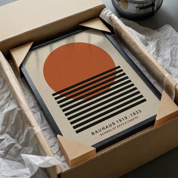 May include: Framed Bauhaus poster with geometric design featuring rust orange semicircle above black and white horizontal stripes on cream background. Print shown in black frame being unpacked from cardboard box with tissue paper wrapping. Modern minimalist artwork styled in packaging presentation with neutral beige tones.