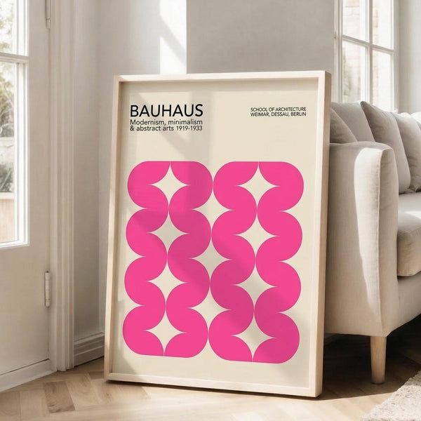 May include: Pink geometric poster in light wooden frame leaning against white wall next to beige armchair. The artwork features bright pink curved shapes forming a repeating pattern on cream background with "BAUHAUS" text at top. Natural lighting from nearby windows creates soft shadows on hardwood floor.