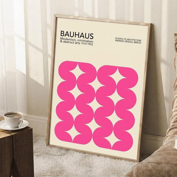May include: Framed poster with cream background featuring bright pink wavy geometric pattern creating interlocking curved shapes. Text at top reads "BAUHAUS Modernism & abstract arts 1919-1933" and "SCHOOL OF ARCHITECTURE WEIMAR, DESSAU, BERLIN". Light wood frame leans against white wall next to wooden side table with white coffee cup, beige knit throw blanket visible on white textured rug.