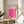 May include: Framed poster with pink geometric wavy pattern on cream background, displayed on wooden round side table next to beige upholstered chair. Natural lighting creates soft shadows across white walls. Green plant leaves visible in corner. Poster features Bauhaus text and repeating curved shapes forming diamond-like negative spaces in bright magenta pink color.