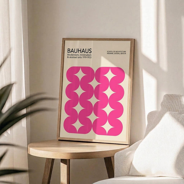 May include: Framed poster with pink geometric wavy pattern on cream background, displayed on wooden round side table next to beige upholstered chair. Natural lighting creates soft shadows across white walls. Green plant leaves visible in corner. Poster features Bauhaus text and repeating curved shapes forming diamond-like negative spaces in bright magenta pink color.