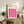 May include: Framed Bauhaus-style poster with bright pink wavy geometric pattern displayed on rustic wooden sideboard in modern living room setting, shown alongside potted fern, ceramic vase, books, and beige upholstered chair with throw pillows.