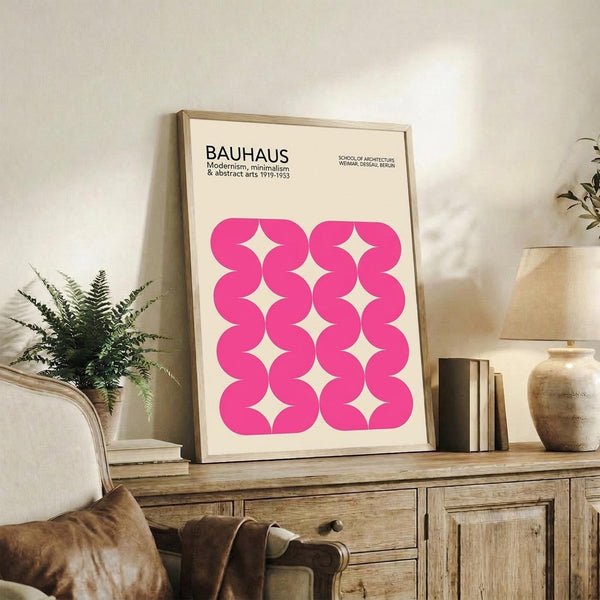 May include: Framed Bauhaus-style poster with bright pink wavy geometric pattern displayed on rustic wooden sideboard in modern living room setting, shown alongside potted fern, ceramic vase, books, and beige upholstered chair with throw pillows.
