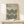 May include: Framed Bauhaus-style poster with geometric black concentric circles pattern on beige background, displayed on light wood console table with small white wrapped gift. Thin natural wood frame, minimalist modern interior setting with white walls and soft natural lighting.
