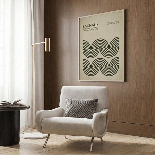 May include: Beige wall art with Bauhaus text and flowing black wave patterns, displayed above a cream upholstered armchair with geometric pillow. Scene features brass floor lamp, dark round side table with open book, wood paneled wall, and sheer curtains. Minimalist modern interior styling with neutral tones and clean lines.