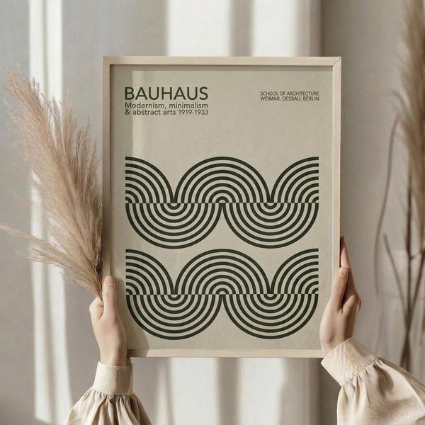 May include: Person in beige sweater holding framed Bauhaus poster featuring black concentric circles and arcs in geometric pattern on cream background. Natural wood frame, dried pampas grass visible, soft neutral lighting near white curtained window.