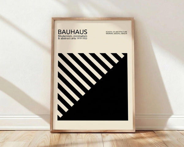 May include: Framed Bauhaus-inspired poster with black and white geometric design featuring diagonal stripes and bold triangle shape, displayed against white wall on wooden floor with natural lighting creating soft shadows across the scene.