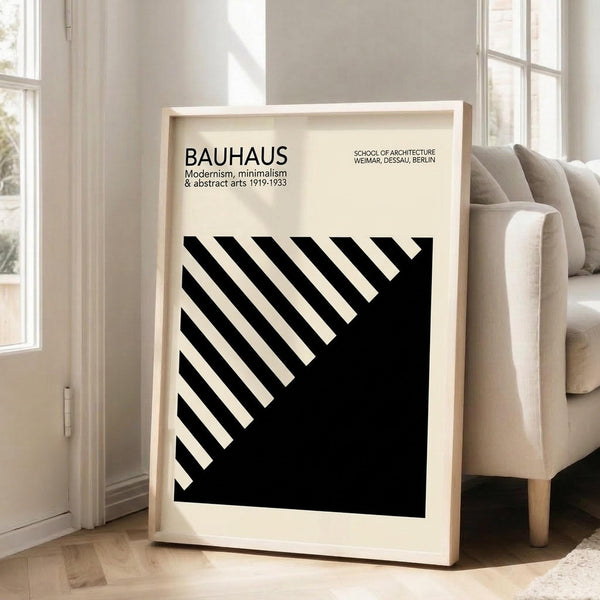 May include: Large framed poster with cream background leaning against white wall next to beige sofa. Black text reads "BAUHAUS Modernism, minimalism & abstract arts 1919-1933" and "SCHOOL OF ARCHITECTURE WEIMAR, DESSAU, BERLIN". Features bold black diagonal striped geometric design in lower portion. Natural wood frame, bright room with windows, hardwood floors.