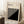 May include: Framed Bauhaus poster with black and white geometric design featuring diagonal stripes and solid black triangle, displayed in light wood frame leaning against white wall corner. Scene shows wooden side table with white coffee cup and saucer, beige textured rug, and soft natural lighting creating shadows on the minimalist interior setting.