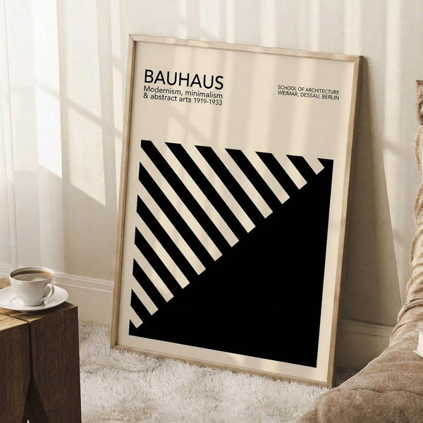 May include: Framed Bauhaus poster with black and white geometric design featuring diagonal stripes and solid black triangle, displayed in light wood frame leaning against white wall corner. Scene shows wooden side table with white coffee cup and saucer, beige textured rug, and soft natural lighting creating shadows on the minimalist interior setting.