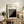 May include: Black and white Bauhaus-style geometric poster with diagonal stripes in a light wood frame, displayed on a rustic wooden sideboard alongside ferns, books, and ceramic pottery in a neutral-toned living room setting with warm natural lighting and textured fabrics.