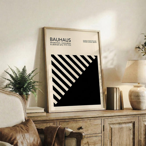 May include: Black and white Bauhaus-style geometric poster with diagonal stripes in a light wood frame, displayed on a rustic wooden sideboard alongside ferns, books, and ceramic pottery in a neutral-toned living room setting with warm natural lighting and textured fabrics.