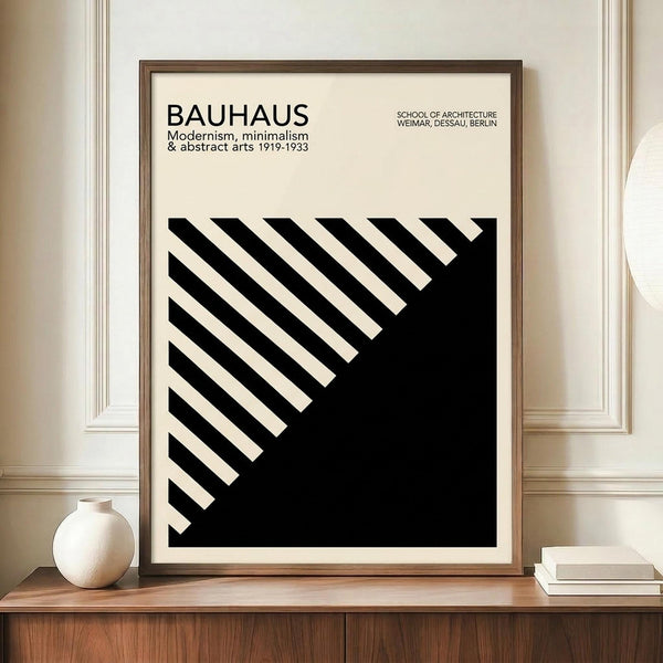 May include: Framed Bauhaus modernist poster with black and white geometric diagonal stripe design displayed on wooden sideboard next to white ceramic vase and books. Features cream background with bold black angular pattern and typography referencing the German design school's history from 1919-1933 across Weimar, Dessau, and Berlin locations.