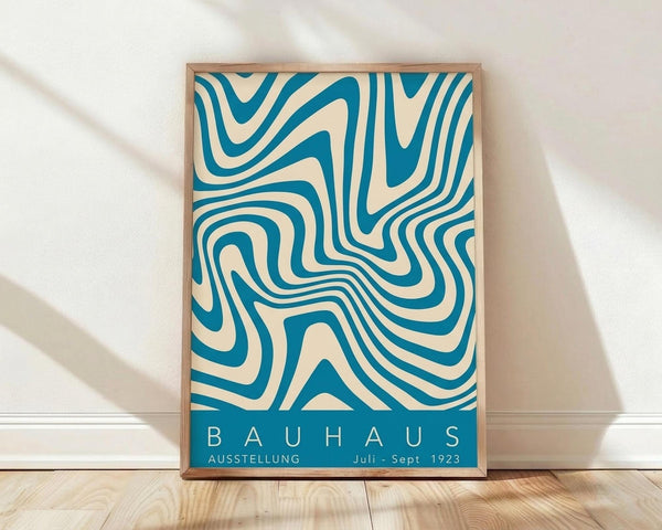 May include: Framed vintage-style poster with flowing teal and cream wavy lines in abstract pattern, displayed against white wall on light wooden floor. Natural light creates soft shadows across the scene. Thin wooden frame surrounds the geometric design with "BAUHAUS" text and German exhibition details at bottom.