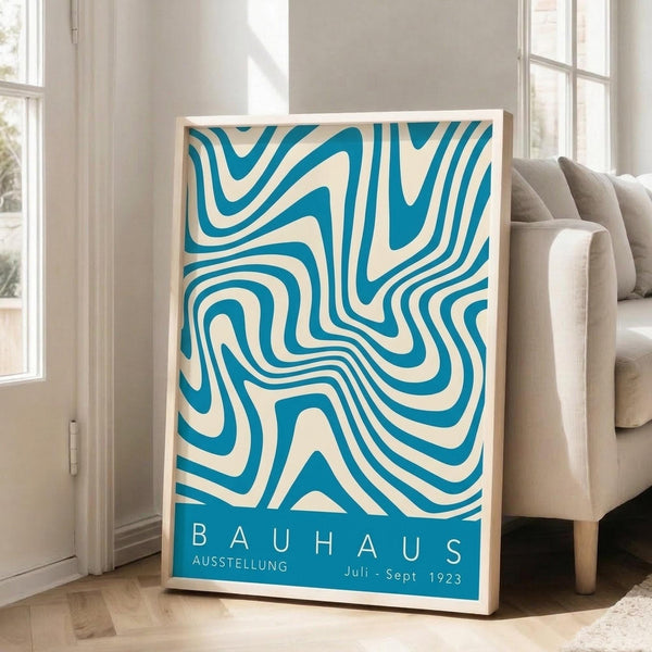 May include: Framed vintage-style Bauhaus exhibition poster with flowing turquoise and cream wavy lines creating an abstract optical pattern. The poster features white text reading "BAUHAUS AUSSTELLUNG Juli-Sept 1923" at bottom. Natural wood frame leans against white wall next to beige upholstered sofa in bright, minimalist living room with tall windows and wooden floors.