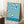 May include: Framed poster with flowing blue and cream swirled pattern leaning against white wall corner. Wooden frame surrounds abstract wavy design with "BAUHAUS AUSSTELLUNG Juli-Sept 1923" text at bottom. White coffee cup on saucer sits on wooden block nearby, with cream textured throw blanket visible. Soft natural lighting creates shadows on wall.