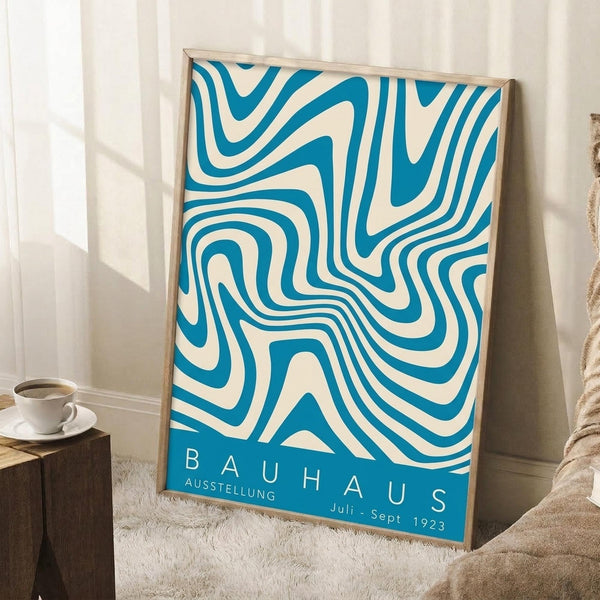 May include: Framed poster with flowing blue and cream swirled pattern leaning against white wall corner. Wooden frame surrounds abstract wavy design with "BAUHAUS AUSSTELLUNG Juli-Sept 1923" text at bottom. White coffee cup on saucer sits on wooden block nearby, with cream textured throw blanket visible. Soft natural lighting creates shadows on wall.