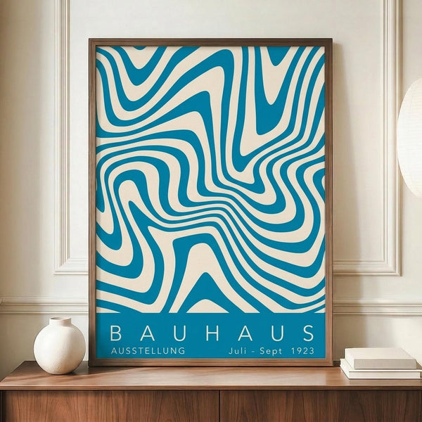 May include: A framed vintage-style exhibition poster with flowing blue and cream wavy lines creating an abstract pattern, displayed on a wooden sideboard against white wainscoted walls. A small white ceramic vase and stack of books sit nearby. The poster features "BAUHAUS" and "AUSSTELLUNG Juli-Sept 1923" text at the bottom in a clean, modernist typeface within a thin wooden frame.