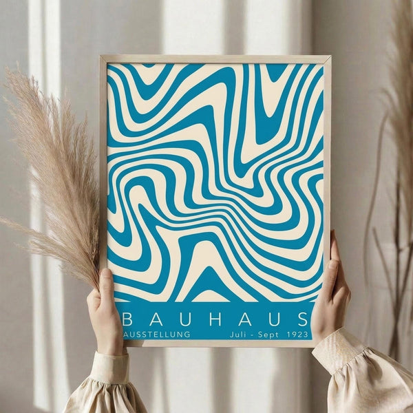 May include: Person holding a framed Bauhaus exhibition poster featuring flowing blue and cream swirled patterns reminiscent of topographic lines. The geometric abstract design fills most of the poster with "BAUHAUS" and "AUSSTELLUNG Juli-Sept 1923" text at bottom. Light wood frame, neutral beige clothing, dried pampas grass visible, soft natural lighting against cream curtained background.