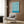 May include: Blue and cream wavy abstract pattern poster mounted on wood paneled wall above beige upholstered armchair with geometric throw pillow. Modern living room setting with brass floor lamp, round black side table with open book, and white curtains. Poster features flowing organic lines in retro mid-century style with exhibition text at bottom.