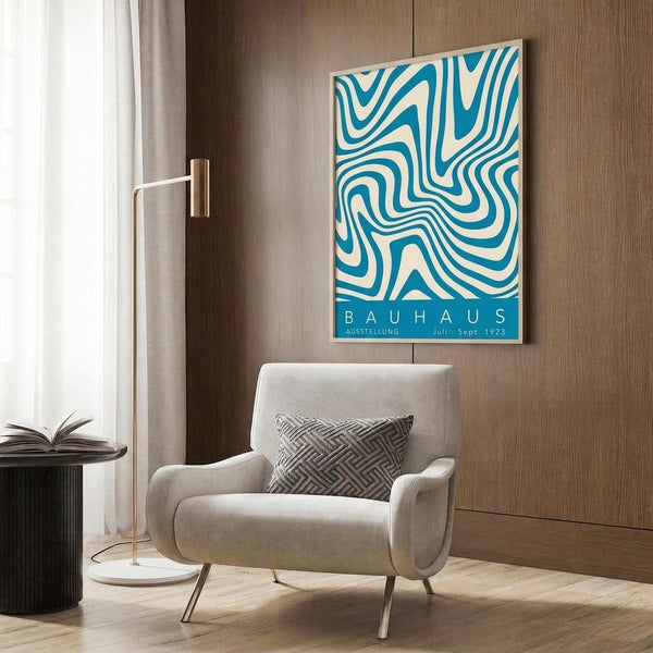 May include: Blue and cream wavy abstract pattern poster mounted on wood paneled wall above beige upholstered armchair with geometric throw pillow. Modern living room setting with brass floor lamp, round black side table with open book, and white curtains. Poster features flowing organic lines in retro mid-century style with exhibition text at bottom.