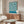May include: Vintage-style Bauhaus exhibition poster with flowing teal and cream swirled pattern displayed above a mid-century modern wooden credenza in a minimalist living room. White sculptural chair and glass coffee table complete the scene with natural lighting and neutral walls.