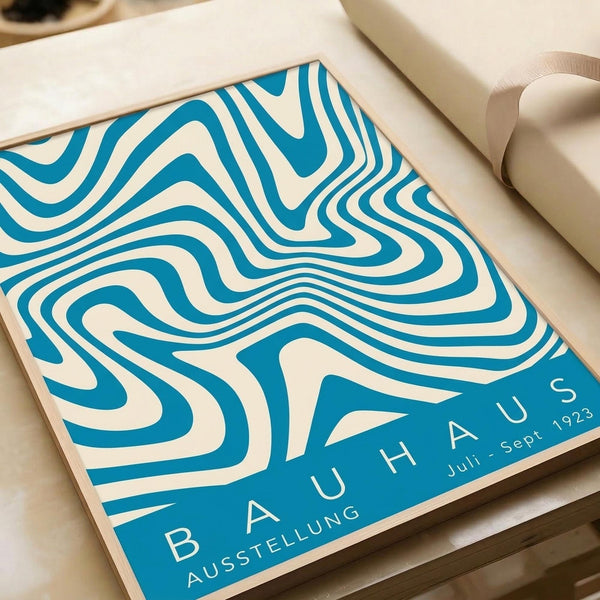 May include: Blue and cream wavy striped abstract poster with flowing, psychedelic-style lines and "BAUHAUS AUSSTELLUNG Juli-Sept 1923" text, displayed in an open book or portfolio on a beige surface next to white paper rolls, showing the vintage exhibition design in warm natural lighting.