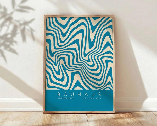 May include: Framed poster with flowing teal and cream wavy lines in a geometric pattern, displayed against white wall on wooden floor. Natural wood frame surrounds the abstract design with "BAUHAUS AUSSTELLUNG Juli - Sept 1923" text at bottom. Plant shadow visible on wall, creating modern minimalist room setting.