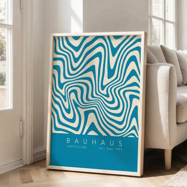 May include: Framed vintage-style poster with blue and cream swirling abstract pattern leaning against white wall next to beige upholstered chair. Natural wood frame, bright sunlight streaming through windows onto hardwood floors in modern minimalist living room setting.