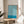 May include: Framed poster with flowing blue and cream wavy lines in psychedelic pattern, displayed on light wood round side table next to beige upholstered chair. Natural sunlight streams across white wall, with plant leaves visible in corner. Thin wood frame surrounds artwork featuring swirling organic shapes and blue text band at bottom.