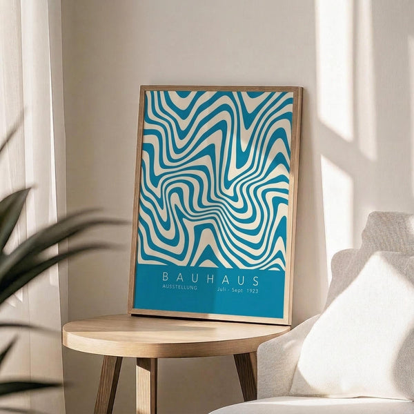 May include: Framed poster with flowing blue and cream wavy lines in psychedelic pattern, displayed on light wood round side table next to beige upholstered chair. Natural sunlight streams across white wall, with plant leaves visible in corner. Thin wood frame surrounds artwork featuring swirling organic shapes and blue text band at bottom.