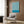 May include: Blue and white swirling abstract pattern poster with "BAUHAUS" text hanging on wood-paneled wall above beige upholstered armchair with geometric patterned pillow. Modern brass floor lamp and dark round side table with open book visible nearby. Warm neutral interior setting with natural lighting from window.
