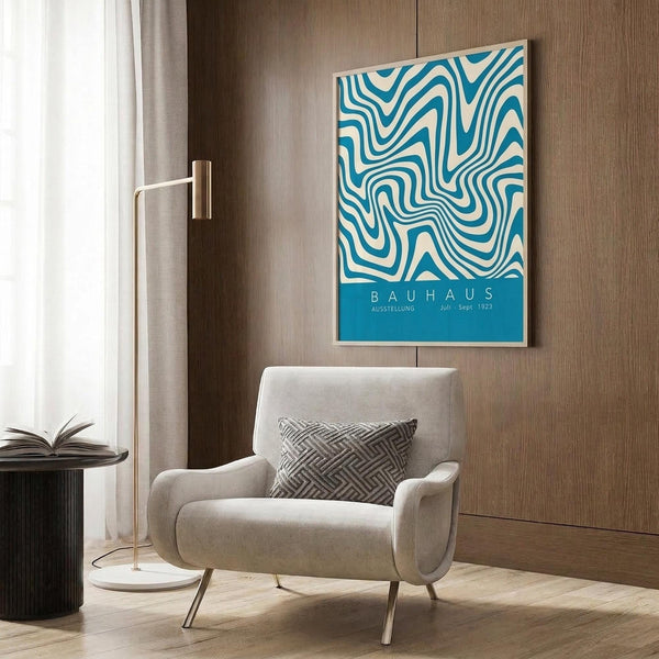 May include: Blue and white swirling abstract pattern poster with "BAUHAUS" text hanging on wood-paneled wall above beige upholstered armchair with geometric patterned pillow. Modern brass floor lamp and dark round side table with open book visible nearby. Warm neutral interior setting with natural lighting from window.