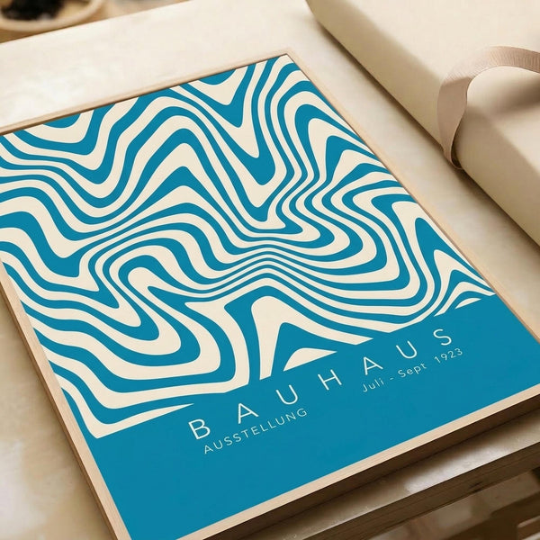May include: Vintage-style poster with flowing turquoise and cream wave pattern displayed on light wood surface next to beige couch. Design features organic curved lines creating topographical effect with "BAUHAUS AUSSTELLUNG Juli-Sept 1923" text in modern typography at bottom right corner.