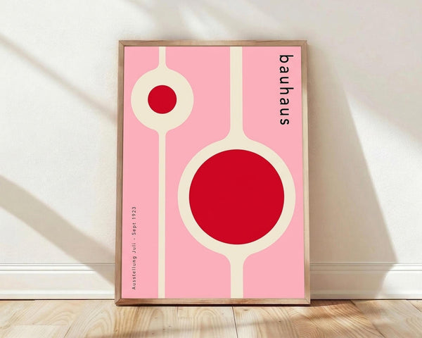 May include: Framed minimalist poster with pink background featuring two connected circular forms in cream and red, displayed against white wall on wooden floor. The geometric design shows Bauhaus-style typography along the right edge. Natural lighting creates soft shadows across the scene.